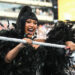 A woman in a black feathered outfit holds a microphone in a wrestling ring, smiling energetically at an event with a large crowd.