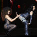Nicki Minaj and Cardi B on stage in revealing outfits, one with curly hair playfully posing behind the other, who has long dark hair.