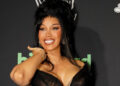 Cardi B smiling at an event, wearing a black dress with her hair styled up. The background features logos and text.