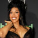 Cardi B smiling at an event, wearing a black dress with her hair styled up. The background features logos and text.