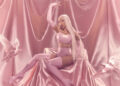 Cardi B with long blonde hair in a pink outfit sits on a silky pink backdrop, surrounded by white doves.