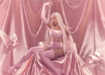 Cardi B with long blonde hair in a pink outfit sits on a silky pink backdrop, surrounded by white doves.