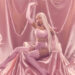Cardi B with long blonde hair in a pink outfit sits on a silky pink backdrop, surrounded by white doves.
