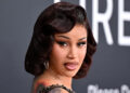Cardi B poses at an event, wearing a glamorous black dress with a vintage hairstyle and visible tattoos.