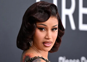Cardi B poses at an event, wearing a glamorous black dress with a vintage hairstyle and visible tattoos.