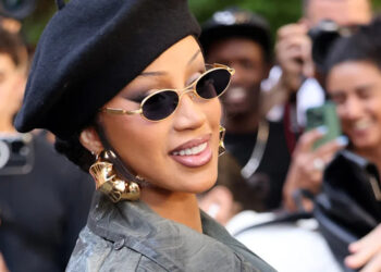 Cardi B in a black beret and sunglasses, smiling at an event. She wears gold earrings and a green coat, with people in the background.