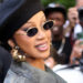Cardi B in a black beret and sunglasses, smiling at an event. She wears gold earrings and a green coat, with people in the background.