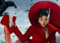 Cardi B in a dramatic red outfit with a large circular headpiece poses with a crow perched on their hand, surrounded by flying birds.