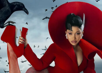 Cardi B in a dramatic red outfit with a large circular headpiece poses with a crow perched on their hand, surrounded by flying birds.
