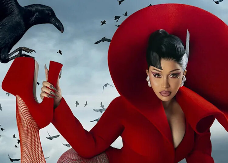 Cardi B in a dramatic red outfit with a large circular headpiece poses with a crow perched on their hand, surrounded by flying birds.