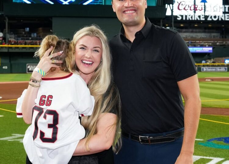 Charlie Kirk smiles with his wife Erika Frantzve, who holds their young daughter in a jersey, while standing on the field at Chase Field.