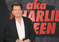 Charlie Sheen at an event, wearing a black jacket and white shirt, standing in front of a backdrop with his name.