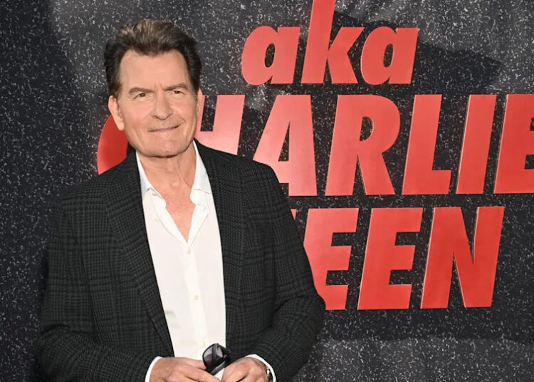 Charlie Sheen at an event, wearing a black jacket and white shirt, standing in front of a backdrop with his name.
