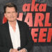 Charlie Sheen at an event, wearing a black jacket and white shirt, standing in front of a backdrop with his name.