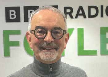 Dónal Doherty pictured wearing dark-rimmed glasses and in a grey zip-up sweater. He is standing in front of BBC Radio Foyle sign.