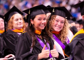 Two smiling graduates in caps and gowns giving thumbs up at a graduation ceremony.