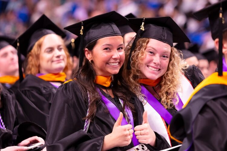 Two smiling graduates in caps and gowns giving thumbs up at a graduation ceremony.