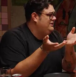 Comedian Caleb Hearon gesturing at a table with his hands, elbows on the table. Caption reads, "I legitimately thought it was funny."