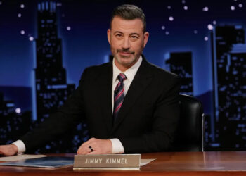 Jimmy Kimmel sitting at a desk on his talk show set, wearing a suit and tie, with a cityscape backdrop.