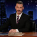 Jimmy Kimmel sitting at a desk on his talk show set, wearing a suit and tie, with a cityscape backdrop.