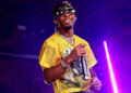 d4vd in a yellow mesh shirt and sunglasses performs on stage, holding a microphone, with purple lighting in the background.