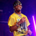 d4vd in a yellow mesh shirt and sunglasses performs on stage, holding a microphone, with purple lighting in the background.
