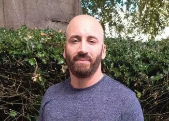 A man in a grey T-Shirt is standing in front of a green hedge. He is bald and has a brown beard. He is smiling at the camera.