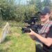 A smiling man in a black cap and T-shirt with a video camera in hand and a checked shirt draped over his shoulder. He stands in a graveyard, filming.