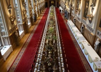 A long banquet table with white place settings and flowers on a red carpet in a lengthy royal hallway lit by candlelight
