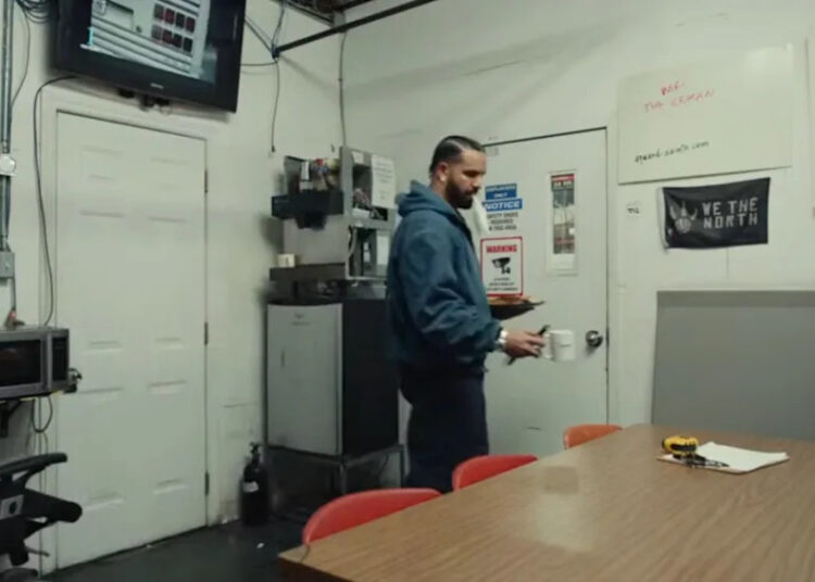 Drake in a blue uniform stands near a wall with a door, bulletin board, and blue bins. The sign above reads