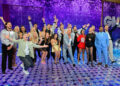 The Dancing With the Stars season 34 cast poses together on the Good Morning America stage, smiling and cheering in front of a sparkly purple backdrop.