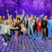 The Dancing With the Stars season 34 cast poses together on the Good Morning America stage, smiling and cheering in front of a sparkly purple backdrop.