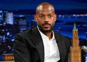 Marlon Wayans in a black suit on a talk show set, cityscape backdrop behind him.