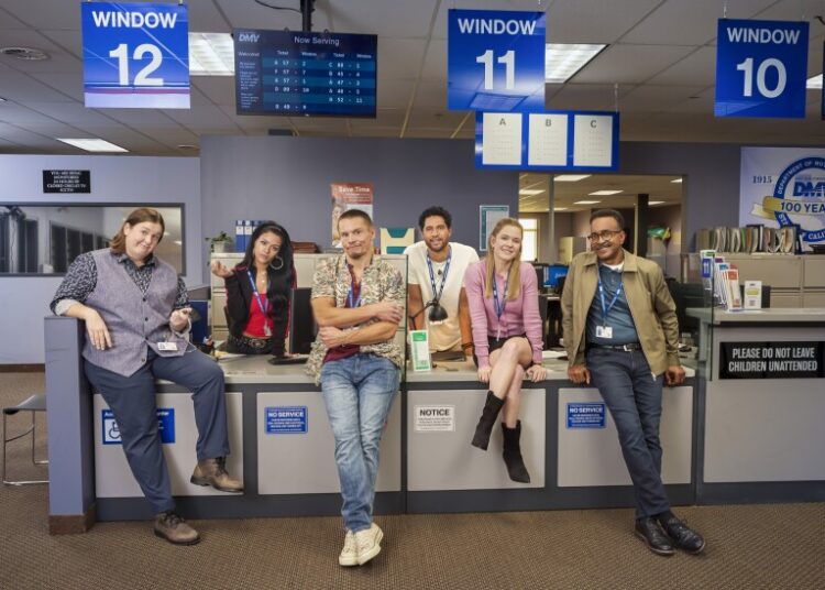 A group of six people - three women and three men - wearing work badges on blue lanyards around their desks stand and sit in front of and behind a desk in an office. A sign partially visible in the back reads “DMV: 100 Years.” A “Now Serving” screen hangs from the ceiling, along with signs that read “Window 12,” “Window 11,” “Window 10.” A sign posted on the desks reads “Please Do Not Leave Children Unattended.”