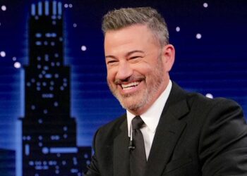 A man in a black suit and tie smiles while sitting at a desk on a talk show set, reminiscent of Jimmy Kimmel, with a city skyline and night sky backdrop behind him.