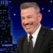 A man in a black suit and tie smiles while sitting at a desk on a talk show set, reminiscent of Jimmy Kimmel, with a city skyline and night sky backdrop behind him.