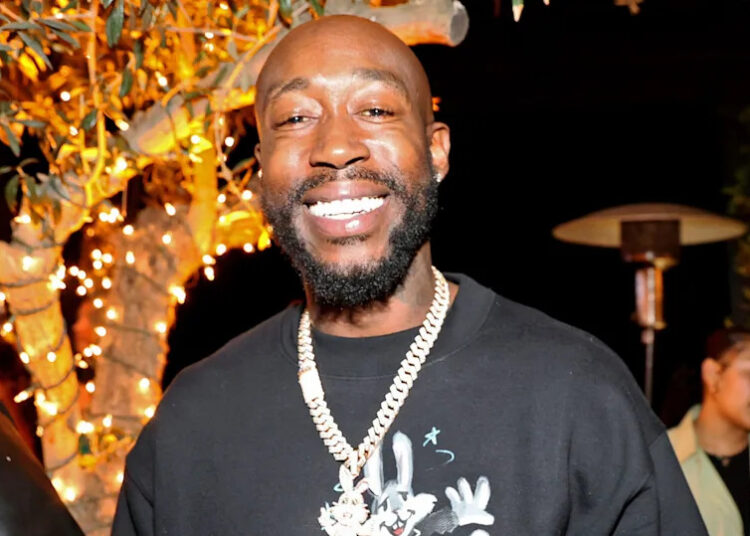 Freddie Gibbs smiling at an event, wearing a black shirt and a gold chain, with decorative lights in the background.