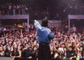 Raf Saperra singing to a large crowd, wearing a blue shirt and black cap, with his left arm outstretched, pointing the black microphone to the crowd. The fans have their hands in the air and are shouting. There are mental stairs and an upper level where fans are also standing.
