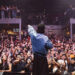 Raf Saperra singing to a large crowd, wearing a blue shirt and black cap, with his left arm outstretched, pointing the black microphone to the crowd. The fans have their hands in the air and are shouting. There are mental stairs and an upper level where fans are also standing.