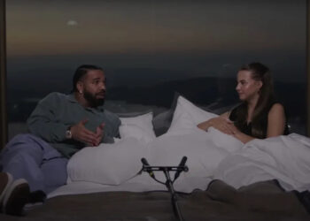 Drake and Bobbi sit on a bed, engaged in conversation. A microphone is in front of them, with a scenic view in the background.