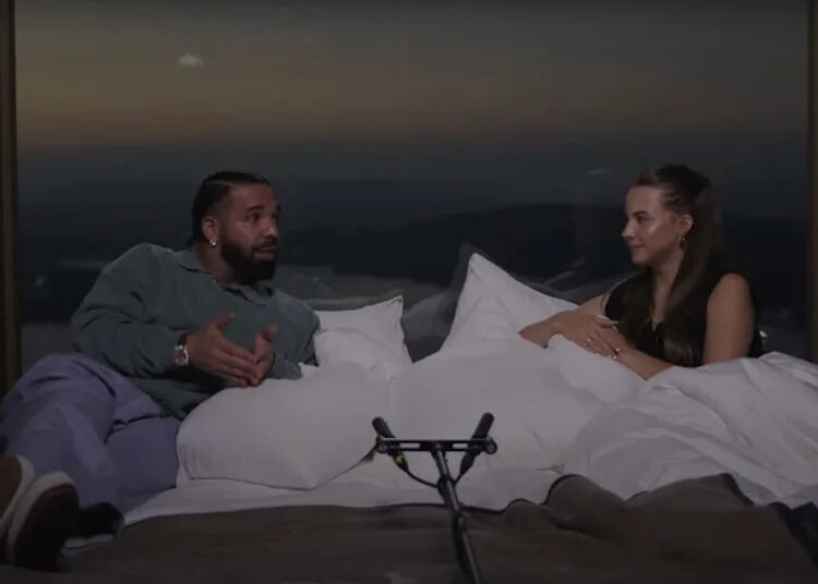 Drake and Bobbi sit on a bed, engaged in conversation. A microphone is in front of them, with a scenic view in the background.