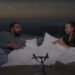 Drake and Bobbi sit on a bed, engaged in conversation. A microphone is in front of them, with a scenic view in the background.