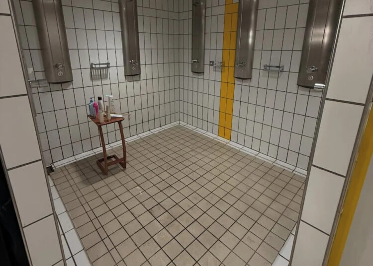 Public shower room with four showerheads, tiled walls, a small stool with toiletries, and a yellow stripe on the wall.