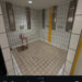 Public shower room with four showerheads, tiled walls, a small stool with toiletries, and a yellow stripe on the wall.