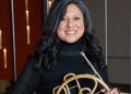 A Latinx woman wears black and holds a French horn.