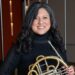 A Latinx woman wears black and holds a French horn.