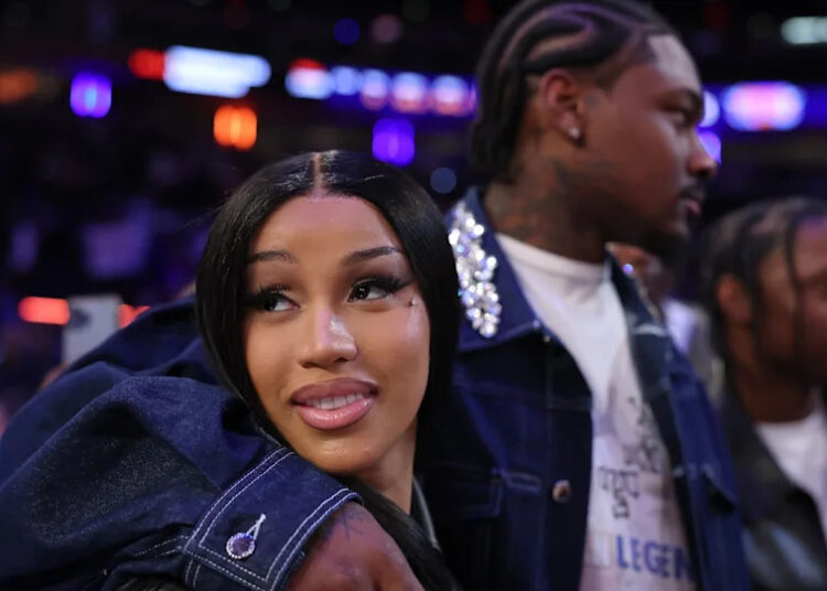 A woman with long black hair is smiling, embraced by a man with braided hair. They're in a dimly lit, colorful venue.
