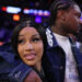 A woman with long black hair is smiling, embraced by a man with braided hair. They're in a dimly lit, colorful venue.