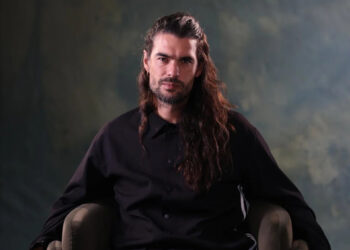 A bearded man with long hair sits in a chair.