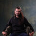 A bearded man with long hair sits in a chair.